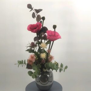 Composition florale artificielle garnie de Pavots roses, de 2 chardons, d'Hamamélis rose et beige, de branches d'Eucalyptus pourpres et vertes. Vase en céramique gris brillant.