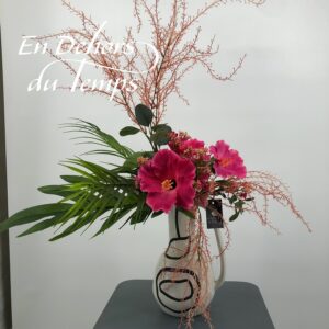 Composition florale artificielle garnie d'hibiscus rose, fleurs de sedum, branchage rose et feuilles de palme. Vase en céramique blanc , anse et motif art contemporain.